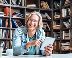 Blonder Student mit Brille sitzt in einer Bibliothek mit einem Tablet in der Hand