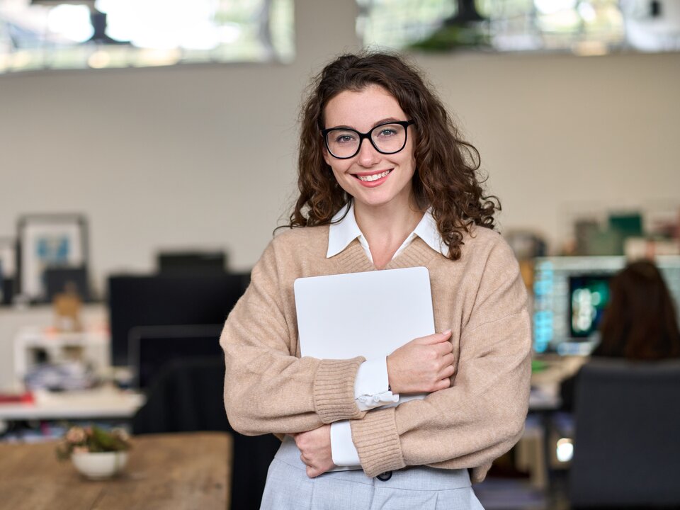 Mädchen mit dunklen Haaren steht in einem Büro und hält einen Laptop in ihren Händen