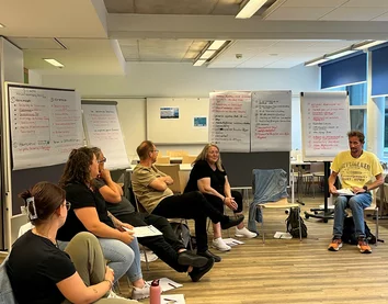 Gruppe von Personen in einem Schulungsraum mit Flipcharts, ein Mann spricht.